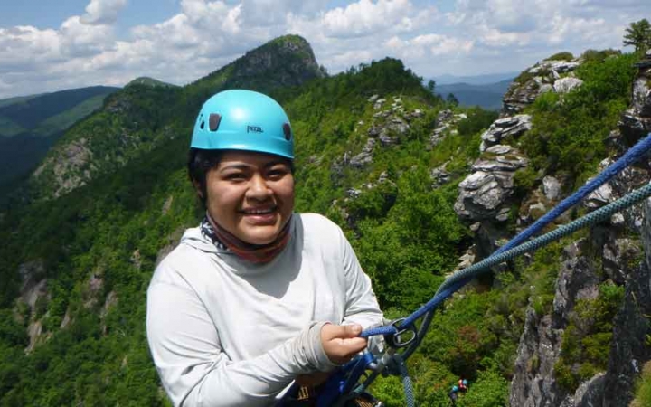 a student pauses to smile at the camera while rock climbing on an outdoor leadership course in the blue ridge mountains. They are wearing safety gear and are secured by ropes.